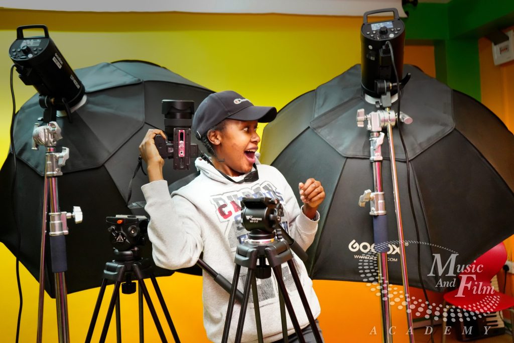 A photography student holds a DSLR camera on a tripod next to professional Godox studio strobes and large black umbrellas during an advanced certificate in photography class at the Music and Film Academy in Nairobi.