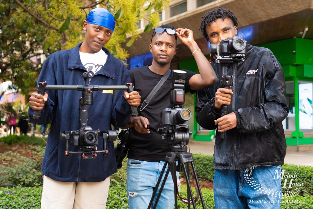 Three videography students operating a dual-handle motorized gimbal stabilizer, a camera tripod with a Godox speedlight, and a single-handed 3-axis electronic stabilizer during an outdoor practical class in Nairobi.