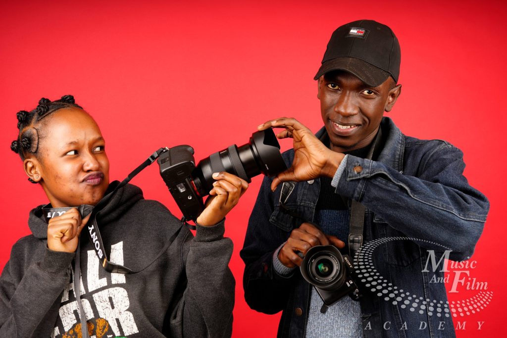 Two photography students adjusting a Sony mirrorless camera lens with a hood against a red background during a Certificate in Photography class in Nairobi.