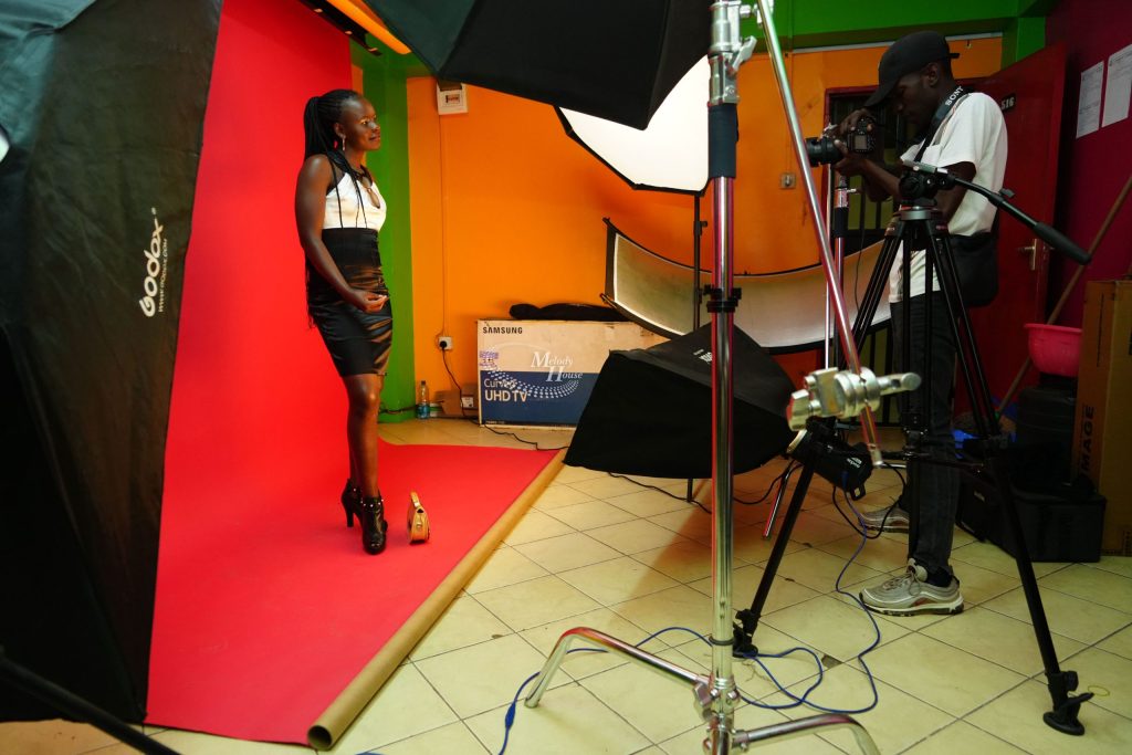 A videography student operating a camera on a professional fluid-head tripod, filming a talent standing against a red backdrop under a continuous Godox softbox lighting array in a studio class in Nairobi.
