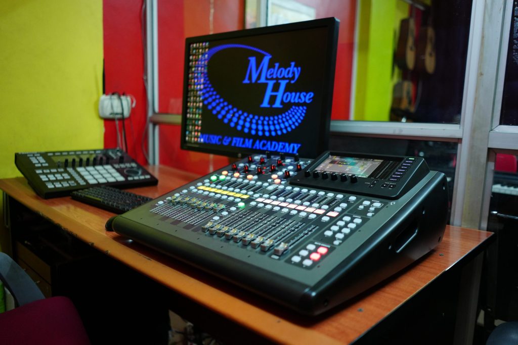A professional digital mixing console with motorized faders, a control monitor displaying the Music and Film Academy logo, a keyboard, and a pad controller inside a recording studio setup in Nairobi.