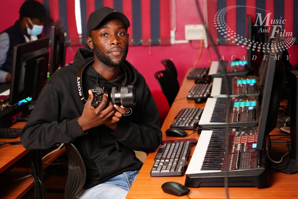 A video editing student wearing a black cap and Vans hoodie holding a Sony mirrorless camera at a high-performance post-production terminal and workstation during a Basic Certificate in Video Editing class in Nairobi.